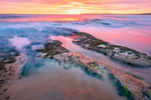 A dramatic sunset over waves crashing into rock pools along a beach - Australian Stock Image