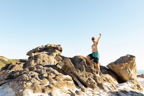 a father wearing board shorts and no shirt standing on top of rocks at a beach - Australian Stock Image