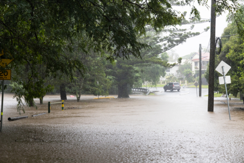 A flooded road and park in a low lying area of Red Hill. A car drives away from floodwater in rain - Australian Stock Image