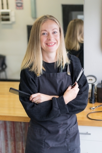 A happy young woman barber holding scissors and a comb and wearing an apron - Australian Stock Image
