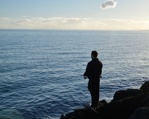 A man fishing alone from some rocks in the bay - Australian Stock Image