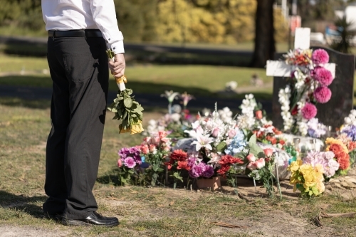 A man in a dress shirt and pants visiting a grave in a cemetery graveyard holding yellow roses - Australian Stock Image