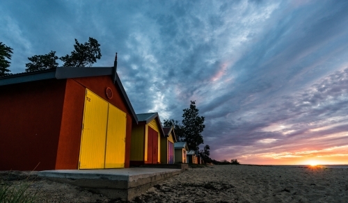 A row of brightly coloured beach huts under a dramatic sky at sunset.