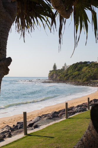 A sandy beach and grass area framed by tree foliage leads to an empty beach with a rocky point break - Australian Stock Image