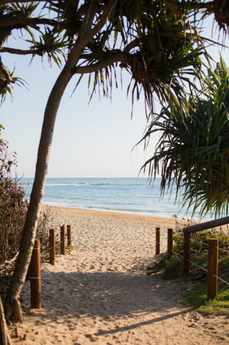 A sandy beach pathway framed by tree foliage leads to an empty beach with gentle waves - Australian Stock Image