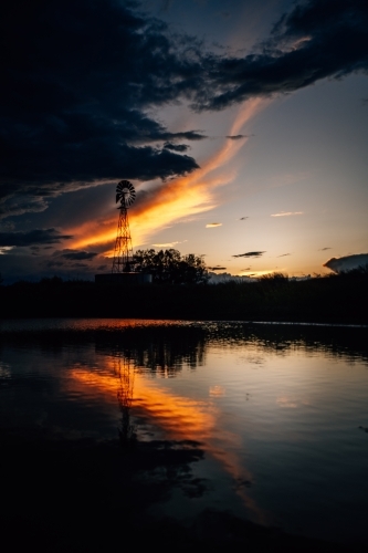 A silhouette of the tall windmill and trees at sunset by dam - Australian Stock Image