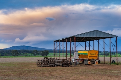 A truck and farm machinery under a hay shed on farmland at sunset