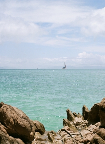 A view from a rocky shore with a clear turquoise water and a sailboat at a distance. - Australian Stock Image