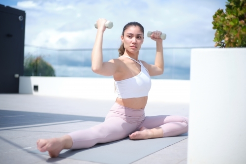 A woman in a white tank top and pink leggings working out using white dumbbells - Australian Stock Image
