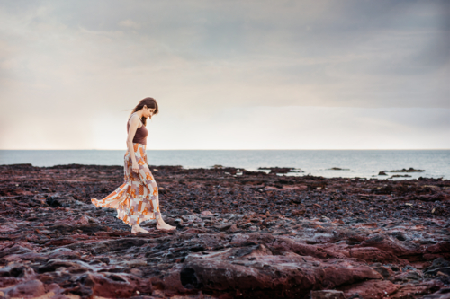 A woman wearing a long skirt walking on a rocky shore with ocean as the background - Australian Stock Image