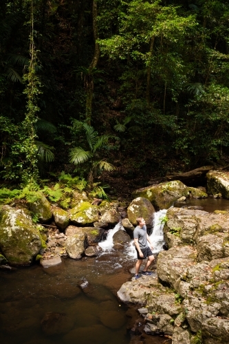 A young male standing beside a small waterfall surrounded by green rainforest.