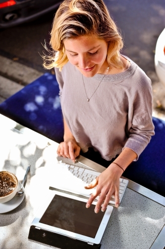 a young woman enjoys her cappuccino whilst working outdoors in a cafe with her tablet device - Australian Stock Image