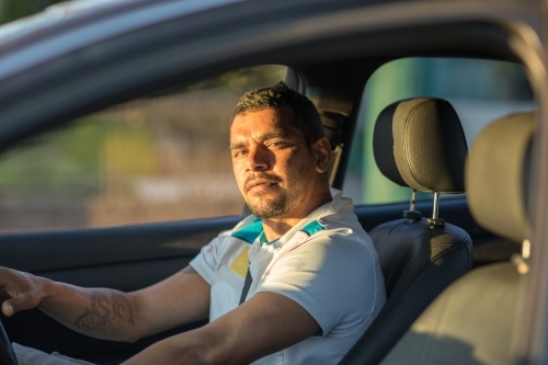 Aboriginal man seen through passenger window driving a car with dark interior - Australian Stock Image