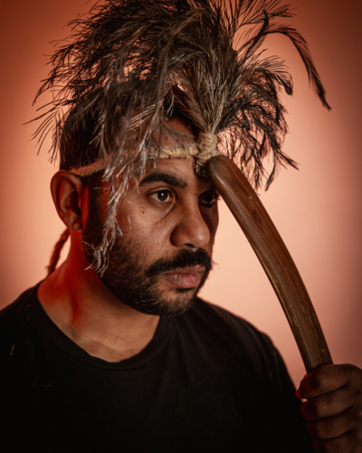 Aboriginal man wearing feathered headdress holding wooden boomerang to his forehead - Australian Stock Image