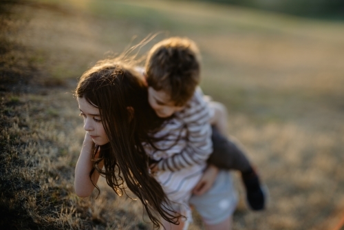 Abstract, blurred image of a sister giving her younger brother a piggyback ride in a field at sunset - Australian Stock Image