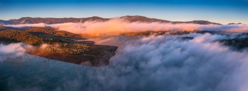 Aerial panorama of early morning light on clouds of fog and hills surrounding a high country lake - Australian Stock Image