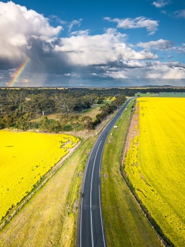 Aerial view of a country road running through farmland with clouds and a rainbow in the distance - Australian Stock Image