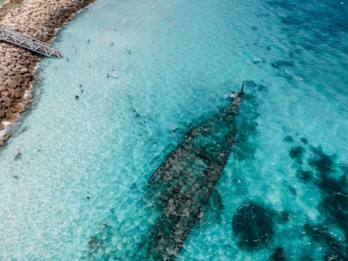 Aerial view of a sunken ship with people swimming, white sand, bushes and grass - Australian Stock Image