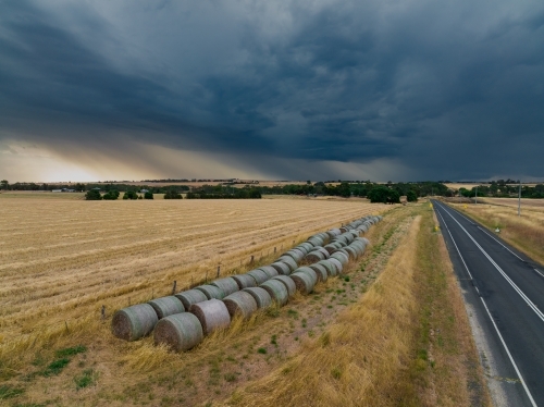 Aerial view of hay bales beside a dry paddock under rain falling from a dark stormy sky
