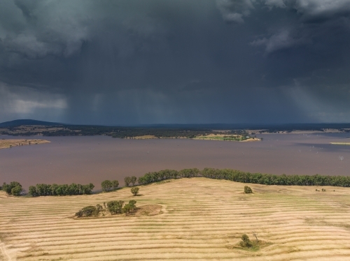 Aerial view of heavy rain falling from storm clouds over a reservoir next to harvested farmland - Australian Stock Image