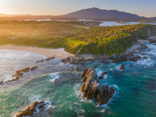 Aerial view of jagged rocks along a rugged coastline at sunset with a mountain in the distance - Australian Stock Image