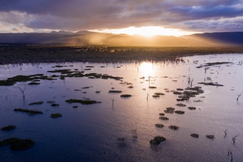 Aerial view of sunset breaking through over a mountain range behind a lake - Australian Stock Image