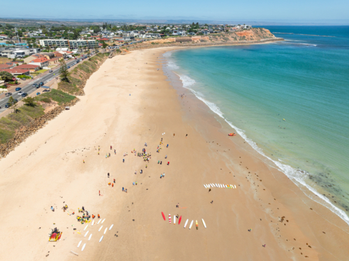 Aerial view of surfboards and beachgoers spread out on a wide sandy beach below a coastal city - Australian Stock Image