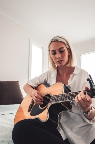 Angled shot of young woman looking down playing guitar on bed - Australian Stock Image