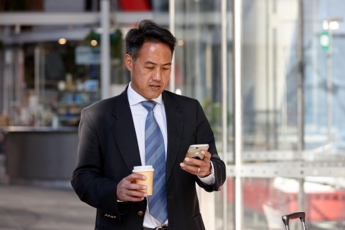 Asian business man checking mobile device outdoors with coffee - Australian Stock Image