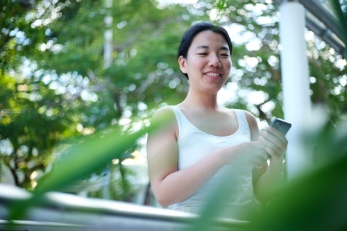 Asian woman on her phone can be seen through the green foliage - Australian Stock Image