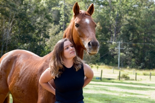 Attractive young woman with long brown hair leaning on chestnut horse - Australian Stock Image