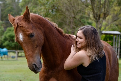 Attractive young woman with long brown hair, smiling and rubbing a chestnut horse - Australian Stock Image