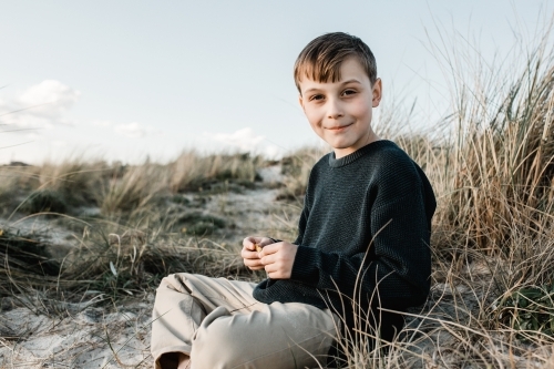 Autistic Boy sitting on a sand dune in the grass looking at the camera - Australian Stock Image