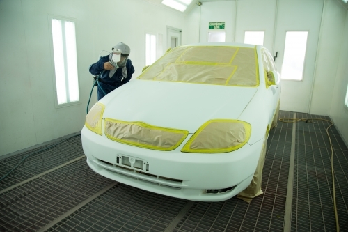 Auto painter in protective suit and mask, spraying a car in painting booth - Australian Stock Image