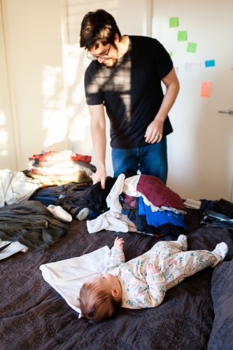 Baby lying on bed in bedroom watching parent fold clean washing - Australian Stock Image