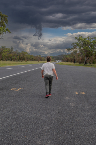 Back of a boy walking away along a road underneath a stormy sky - Australian Stock Image