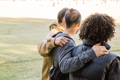 back view shot of three men one curly hair and one with a manbun with arms over each other's back - Australian Stock Image