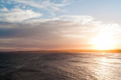 Big, blue, smooth ocean surrounds distant land on the horizon at sunset - Australian Stock Image