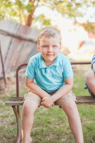 Blond blue eyed 5 year old boy sitting on outdoor bench - Australian Stock Image