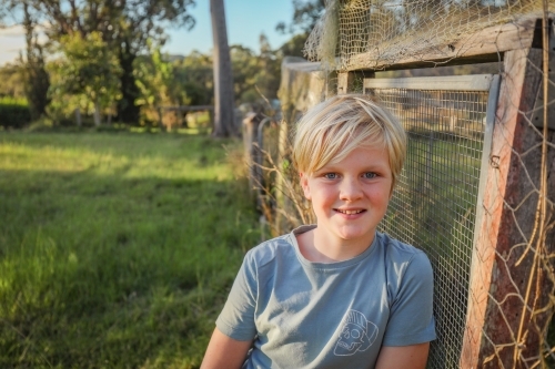 Blonde 10 year old boy relaxed and smiling on farm leaning against chicken coop - Australian Stock Image