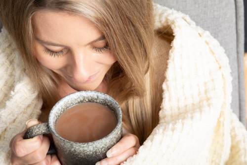 Blonde young lady wrapped in cream blanket hugging a mug - Australian Stock Image