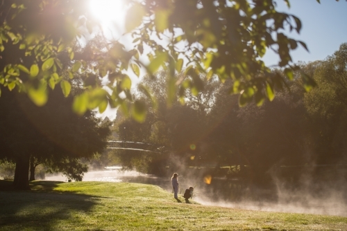 Boy and girl rugged up on cold morning fog day watching ducks in river - Australian Stock Image