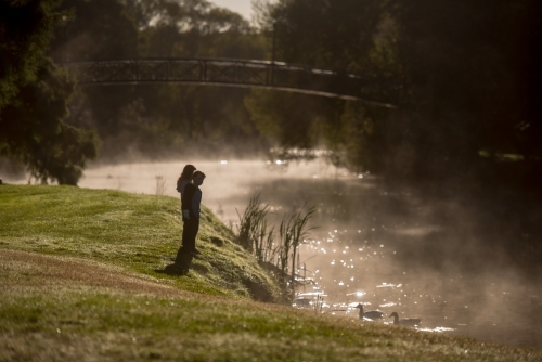 Boy and girl rugged up on cold morning fog day watching ducks in river - Australian Stock Image