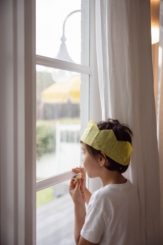 Boy at window wearing a Christmas crown and eating a mince pie - Australian Stock Image