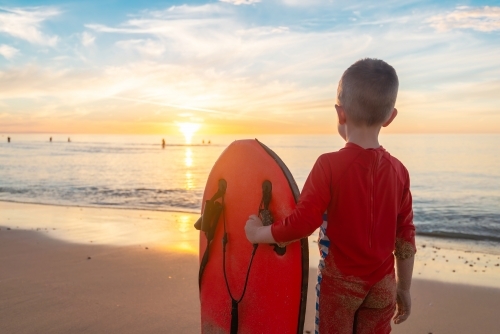 Boy holding bodyboard whilst standing at the beach and watching sunset, Chrieties Beach, SA - Australian Stock Image