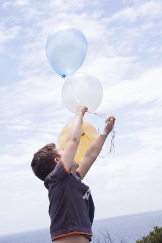 Boy holding bunch of coloured balloons looking up at them - Australian Stock Image