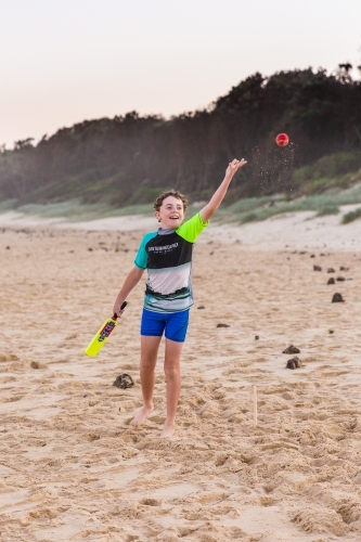 Boy holding cricket bat throwing ball happy having fun on sand at beach - Australian Stock Image