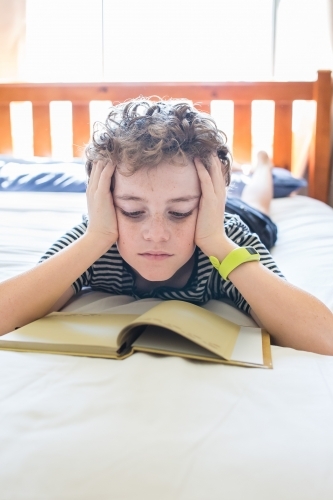 Boy lying on stomach on bed head resting in hands reading book natural light - Australian Stock Image