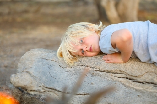 Boy relaxing on log near camp fire - Australian Stock Image