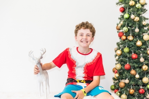 Boy sitting next to Christmas tree holding reindeer smiling excited - Australian Stock Image